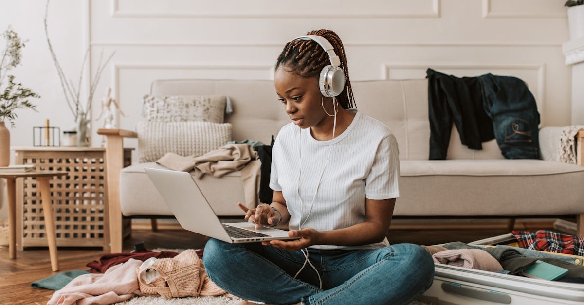 A young woman sits on the floor using a laptop and headphones, amidst clothes and a suitcase.